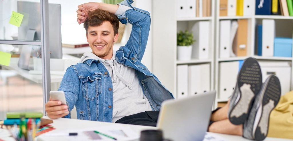 A man in a jean jacket relaxing with his feet up on a desk, looking at his mobile phone