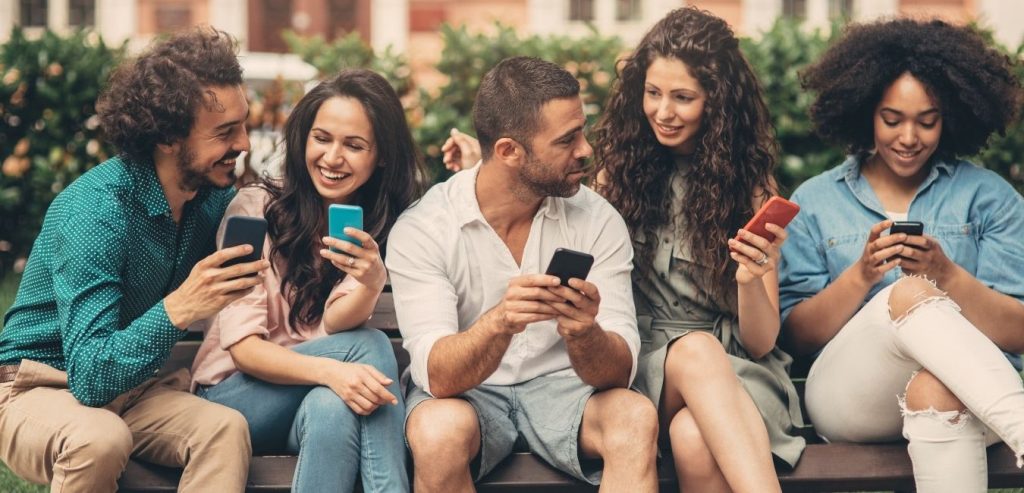 Friends sitting on a park bench having a good time while looking at their mobile phones.